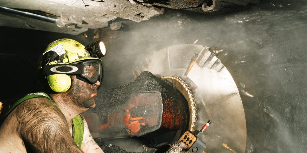 Miner wearing PPE (ear defenders, hard hat and goggles) operating a large coal cutting machine